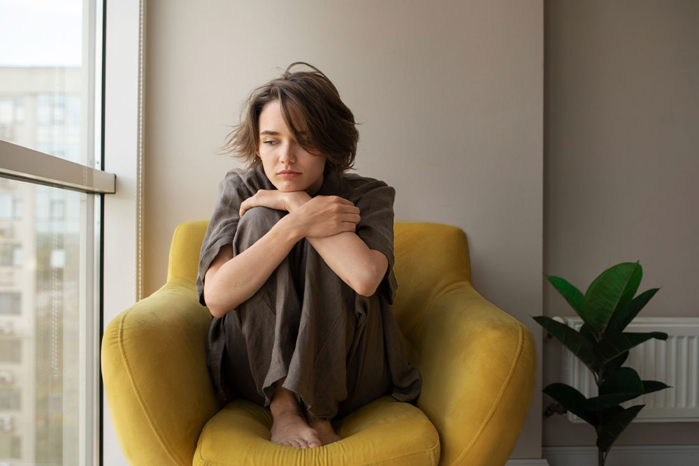 A young woman sitting cross-legged on a bed, holding her head in her knees while sitting